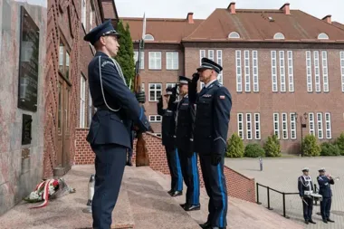 Funkcjonariusze polskiej policji w mundurach galowych stoją na warcie i salutują przed tablicą pamiątkową na budynku z czerwonej cegły.