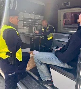 Policjanci w kamizelkach odblaskowych przeprowadzają czynności wewnątrz policyjnego busa z jednym z uczestników zdarzenia.