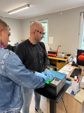 Funkcjonariusz w rękawiczkach pobiera odciski palców mężczyźnie za pomocą skanera biometrycznego w pomieszczeniu biurowym.