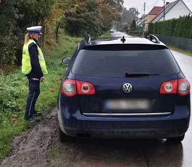 Policjantka w kamizelce odblaskowej stoi przy zaparkowanym na poboczu granatowym volkswagenie passacie na wiejskiej drodze.