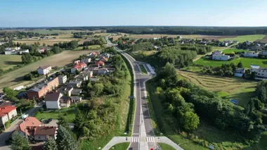 Widok z lotu ptaka na nowo wybudowaną lub wyremontowaną drogę biegnącą przez wiejską, zieloną okolicę z domami jednorodzinnymi i polami.