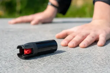 Dłonie osoby leżącej na szarej nawierzchni obok czarnego pojemnika z czerwoną końcówką przypominającego gaz pieprzowy.