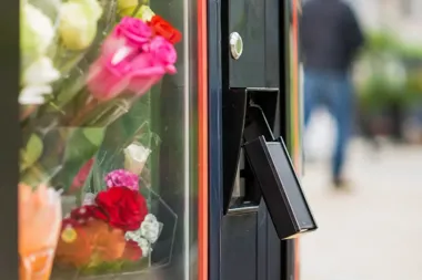 Fragment automatu z bukietami kwiatów za szybą, z wysuniętym czarnym panelem do płatności, na tle rozmytej postaci przechodnia na ulicy.