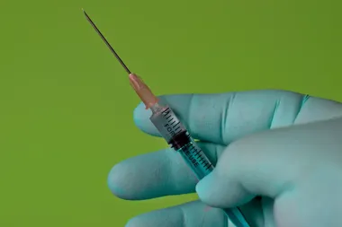 Ręka w niebieskiej rękawicy medycznej trzyma strzykawkę z igłą na jednolitym zielonym tle.