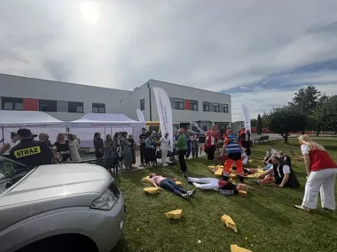Pokaz ratownictwa medycznego na trawie przed budynkiem, z udziałem straży, ratowników, symulowanymi poszkodowanymi i grupą obserwatorów.
